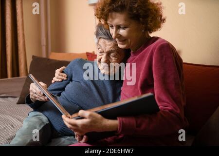 Positive aged ladies looking album photos sitting sofa at home, cheerful friends. Senior woman and her mature nurse watching photo album. Granny showi Stock Photo