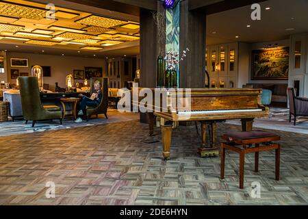 A Steinway grand piano stands at the centre of a lounge at the five-star Dunloe Hotel & Gardens near Killarney, County Kerry, Ireland, beneath a coffered ceiling. In the background, journalist Angela Berg reads in an armchair. Stock Photo