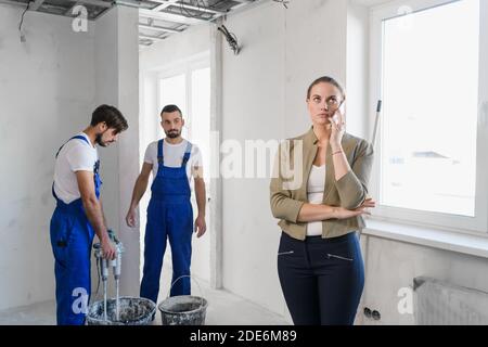 Female inspector talking on a cell phone. Two builders in overalls doing repairs Stock Photo