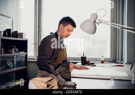 A young shoemaker makes a drawing for a pattern for leather shoes on a table in his workshop. Stock Photo