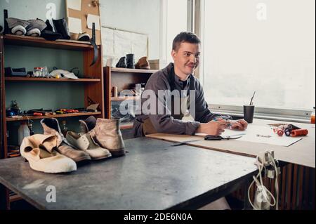 A young shoemaker makes a drawing for a pattern for leather shoes on a table in his workshop. Stock Photo