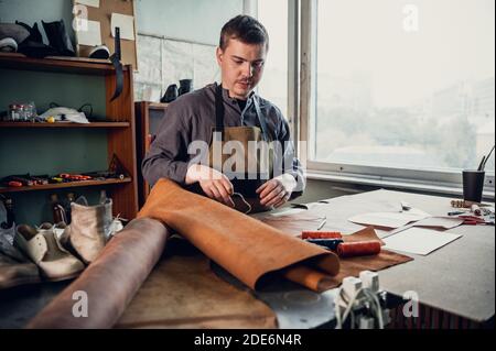A young apprentice in a boot workshop prepares leather for further use on a large table. Stock Photo
