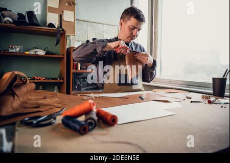 A young tanner makes blanks from leather for shoes at the table, on the table are threads and scissors. Stock Photo