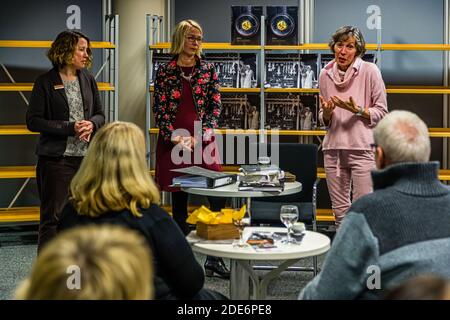 Three women stand in front of a bookshelf with framed black-and-white photographs during a culinary reading at the Neuss city library, Germany, while listeners in the foreground have taken their seats. Several hardcover books lie ready on a round standing table. Stock Photo
