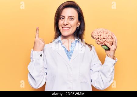 Young beautiful woman wearing doctor coat holding brain smiling with an idea or question pointing finger with happy face, number one Stock Photo