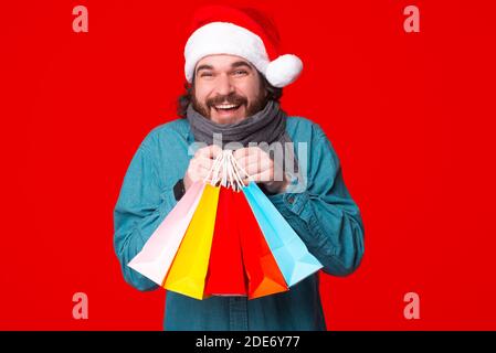 Bearded man holding shopping bags wearing santa hat standong over red background. Stock Photo