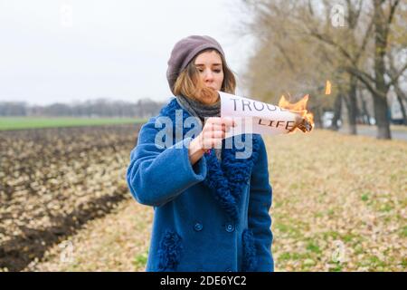 Beautiful young woman in blue coat stay at open area and hold burning paper with printed words Trouble life Stock Photo