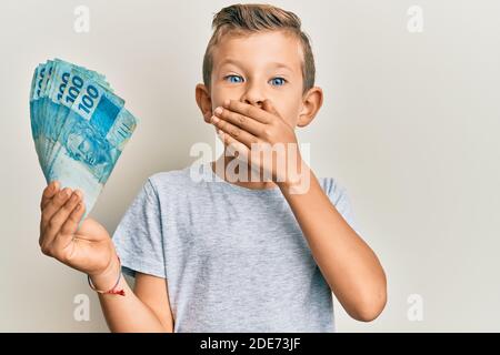 Adorable caucasian kid holding 100 brazilian real banknotes covering mouth with hand, shocked and afraid for mistake. surprised expression Stock Photo