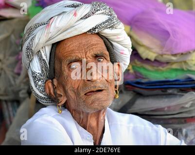 Frail old toothless Indian Rabari man with golden tribal earrings and wrinkled face wears a traditional turban and looks at the viewer. Stock Photo