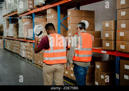 Two male workers lifting hardhats off their heads walking past packages in warehouse  Stock Photo