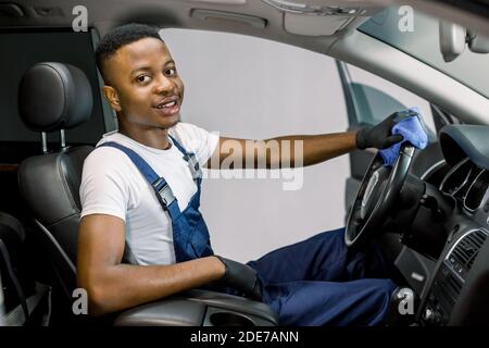 Handsome African young man in uniform and protective gloves, cleaning car interior and steering wheel using microfiber cloth, smiling at camera. Car Stock Photo