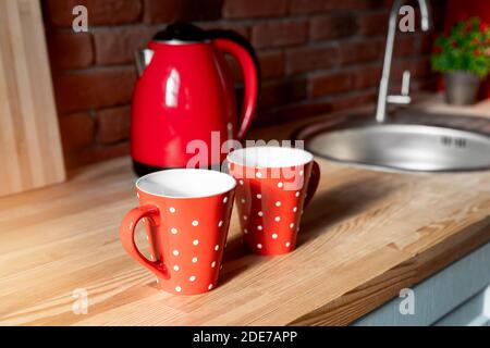 Pair Red polka dot retro vintage coffe tea mugs on wooden kithen counter at rustic room interior on breakfast time. Two hot drink cups and red kettle Stock Photo
