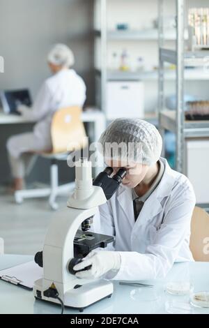 Vertical portrait of young female scientist looking in microscope while studying plant samples in biotechnology lab Stock Photo