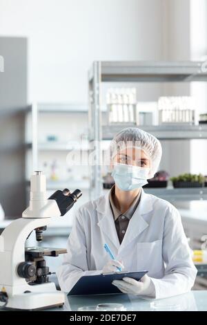Vertical portrait of young female scientist wearing face mask and looking at camera while working in laboratory, copy space above Stock Photo