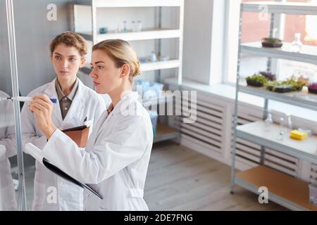 Side view portrait of two female scientists writing on glass wall and holding clipboard while doing research in medical laboratory, copy space Stock Photo