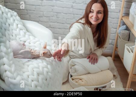 Woman in robe is sitting near wooden shelving, organizing closet by sorting and folding bath towels. Light pink colored Don Sphynx cat Stock Photo