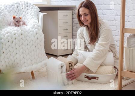 Woman in robe is sitting near wooden shelving, organizing closet by sorting and folding bath towels. Light pink colored Don Sphynx cat Stock Photo