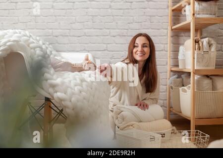 Woman in robe is sitting near wooden shelving, organizing closet by sorting and folding bath towels. Light pink colored Don Sphynx cat Stock Photo