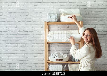 Young woman in warm robe is organizing linen closet with neatly folded towels, sheets and blankets. Stock Photo