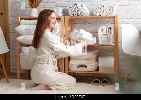 Young woman in warm robe is organizing linen closet with neatly folded towels, sheets and blankets. Stock Photo