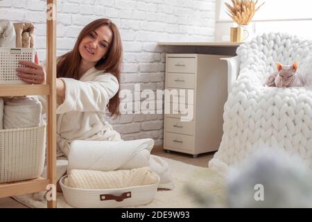 Woman in robe is sitting near wooden shelving, organizing closet by sorting and folding bath towels. Light pink colored Don Sphynx cat Stock Photo