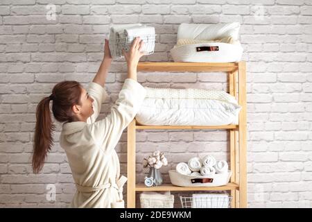Young woman in warm robe is organizing linen closet with neatly folded towels, sheets and blankets. Stock Photo