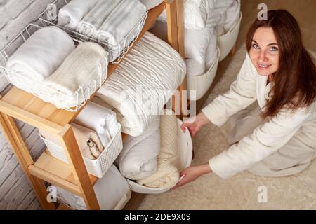 Young woman in warm robe is organizing linen closet with neatly folded towels, sheets and blankets. Stock Photo