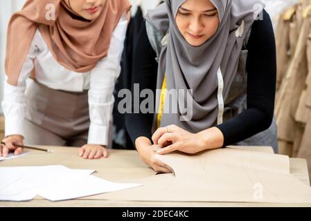 Muslim woman fashion designer working together with her colleague cutting clothing pattern in tailor shop Stock Photo