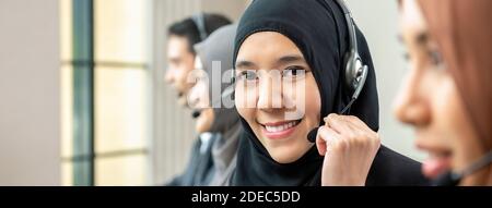Friendly beautiful Asian muslim woman wearing microphone headset working as customer care operator with team in call center office, panoramic web bann Stock Photo