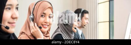 Friendly beautiful Asian muslim woman wearing microphone headset working as customer care operator with team in call center office, panoramic web bann Stock Photo