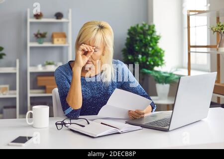 Upset senior woman sitting at desk, holding bankruptcy or debt notification from bank Stock Photo