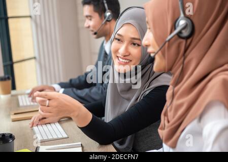 Happy beautiful Asian muslim women working in call center office together with colleagues Stock Photo