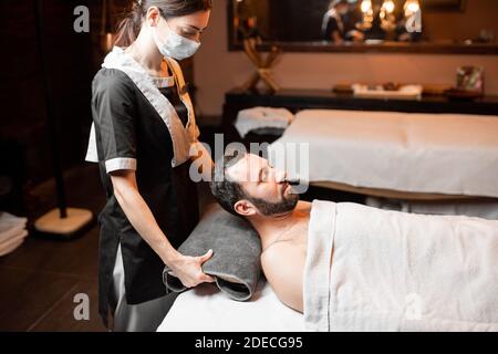 Professional female masseur in medical mask doing facial massage to a male client at Spa salon. Business during the epidemic concept Stock Photo