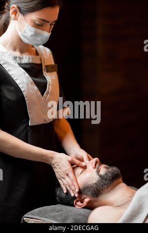 Professional female masseur in medical mask doing facial massage to a male client at Spa salon. Business during the epidemic concept Stock Photo