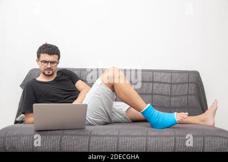 Man with glasses with broken leg in blue splint for treatment of injuries from ankle sprain working on a laptop on couch at home. Stock Photo