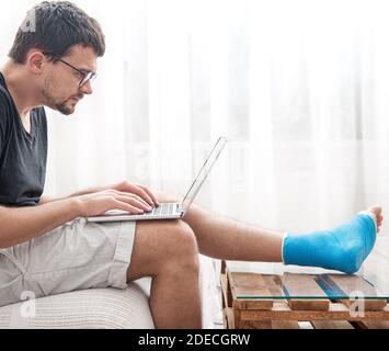 Young man with a broken leg in blue splint for treatment of injurie and ankle sprain is using a laptop at home. Stock Photo