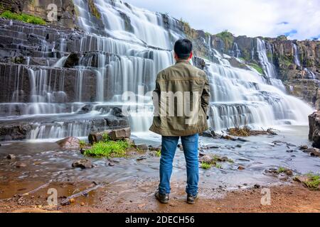 Traveler Man watching beautiful waterfall majestic scenery as a way to relax the soul of his trip around the country in Dalat, Vietnam. Stock Photo