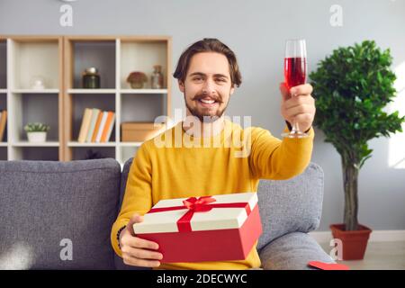 Hipster man sits with a gift and raises a glass of red wine for a birthday or Valentine's Day. Stock Photo