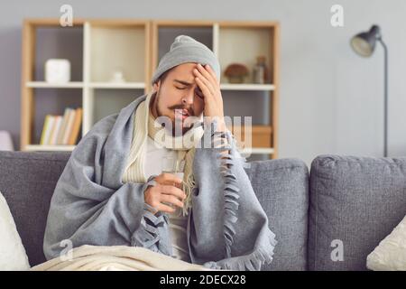 Sick man suffering from a severe headache sits with his eyes closed in pain wrapped in a plaid. Stock Photo