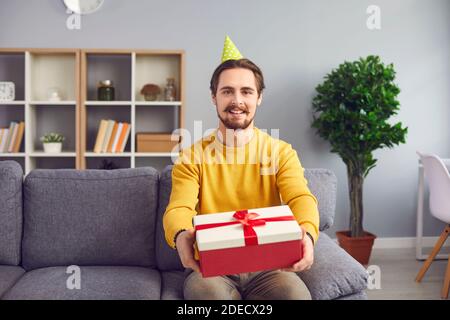 Young positive man in festive clown hat sitting on sofa and showing holiday present box Stock Photo