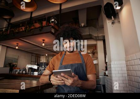 Young male waiter wearing apron uniform in cafe using digital tablet to watch recipe Stock Photo