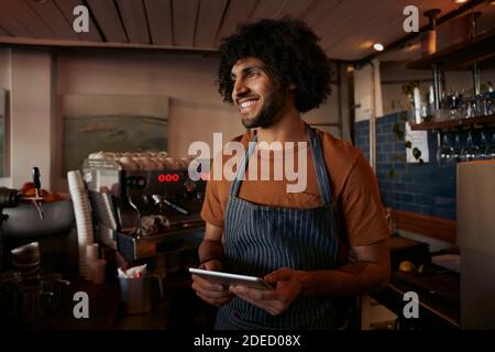 Successful small male business owner wearing apron using digital tablet and looking away Stock Photo