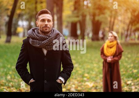 Man stands in a coat and a scarf in the park waiting for his girlfriend or wife who stands behind him or looks like him from the back. Young couple Stock Photo