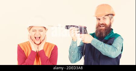 Builder, repairman makes hole in female head. Woman on scared face in helmet, hard hat. Husband annoying his wife. Annoying repair concept. Man with happy face drills head of woman, white background. Stock Photo