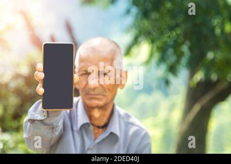 Rural farmer of Indian ethnicity holding blank mobile phone in his hand portrait near green field. focus on mobile screen Stock Photo
