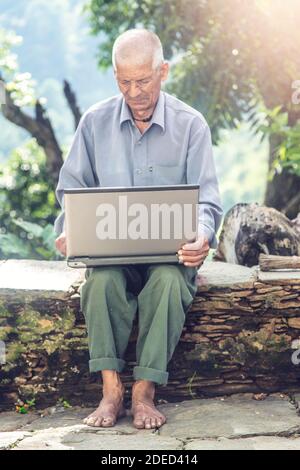 Indian old farmer smiles using laptop and internet. Farmer loves technology, Old farmer works on Laptop. Indian sitting in front of his house Stock Photo