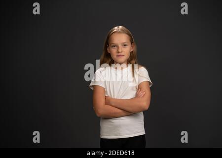 Little girl of 8,10 years with hands folded looks looks questioningly at the camera wearing white t shirt isolated on dark grey or black background Stock Photo