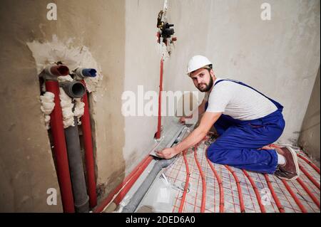Portrait of bearded young man in safety helmet installing pipes in apartment. Male plumber in work overalls installing water trap in the shower. Concept of home renovation and plumbing works. Stock Photo