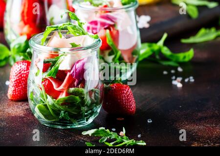 Fresh salad with spicy arugula, goat cheese, strawberry and prosciutto or ham in modern glass jars on black table, selective focus Stock Photo