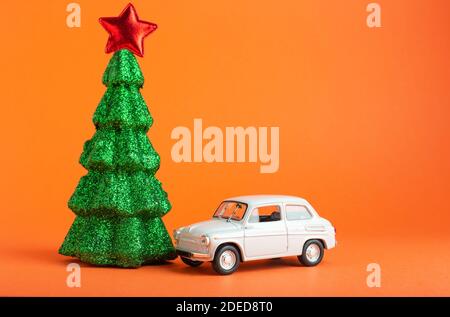 Christmas New Year tree with red star on top near white car toy. Creative miniature Xmas tree and car on Orange color background.. Gift delivery Stock Photo